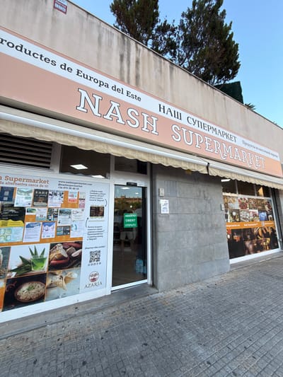 The image shows the storefront of "NASH SUPERMARKET" in Lloret de Mar, Spain. The facade is painted a light peach color, with the store name prominently displayed in large, stylized white letters. Above the store name, there is text in what appears to be Russian, reading "НАШ СУПЕРМАРКЕТ" (Our Supermarket), and to the right of that, "Продукты из восточной европы" (Products from Eastern Europe). Below this, the store's name is written again, this time with the word "SUPERMARKET" in English.
The storefront features a large display window with various advertisements and images of food products, including ice cream, meats, and produce. To the left of the entrance, a wall is covered with numerous flyers and posters. The entrance itself has a green sign that reads "ABIERTO OBERT ОТКРЫТО" (Open in Spanish, Catalan, and Russian).
The sidewalk in front of the store is paved with grey square tiles. A tree with dark green foliage is visible above the building, and the sky is a clear, pale blue. The overall scene depicts a typical street-level view of a small business.