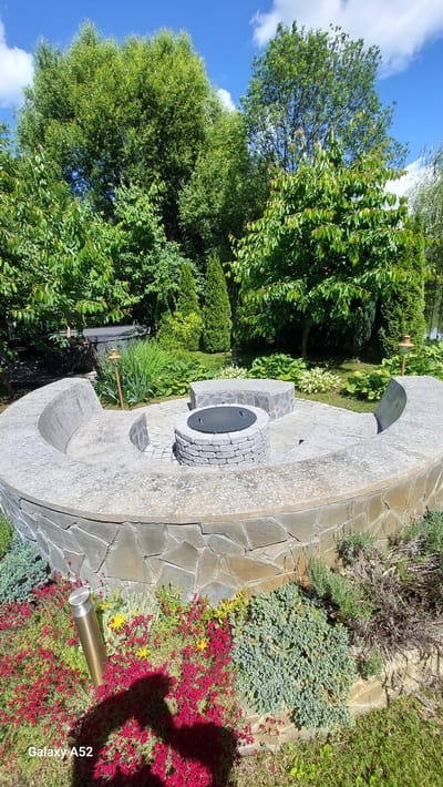 A sunlit, outdoor scene in Zhavoronki, Russia, features a circular stone fire pit area designed for social gatherings. The fire pit is centrally located, constructed with stacked stone rings and a dark metal lid. Surrounding the pit is a curved stone bench, also built with interlocking stones, forming a semi-enclosed seating area. In front of the bench, a low border of red and pink flowers, interspersed with low-lying green and blue-green bushes, adds a touch of vibrant color to the foreground. Two cylindrical metal lamps are visible, one in the floral bed and another partially obscured by greenery near the back of the fire pit. The background is dominated by lush, mature trees and manicured greenery, suggesting a well-maintained garden or park. The sky above is a clear blue with scattered white clouds. A dark-colored hot tub is partially visible to the left, behind the seating area. The overall impression is one of a tranquil, established outdoor living space.