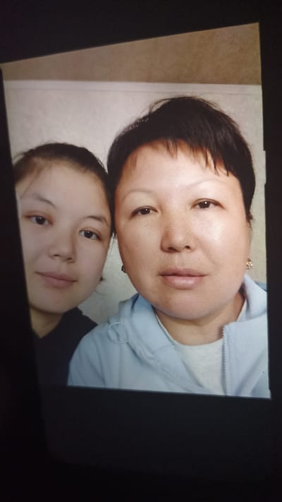 This is a selfie featuring two individuals, likely mother and daughter, positioned close together and looking directly at the camera. The person on the left, a younger woman with dark hair pulled back, is looking slightly off to the side. The person on the right, an older woman with short dark hair, is looking directly at the camera. Both subjects appear to be indoors, with a plain, textured wall visible behind them. The older woman is wearing a light blue zippered jacket over a lighter grey t-shirt. The younger woman is wearing a dark top. Both subjects appear calm and neutral in their expressions. There is no visible text in the image, and no information about the time of day or weather can be inferred from the scene. The setting appears to be a domestic interior, consistent with the provided location context of Samarkandek, Kyrgyzstan.