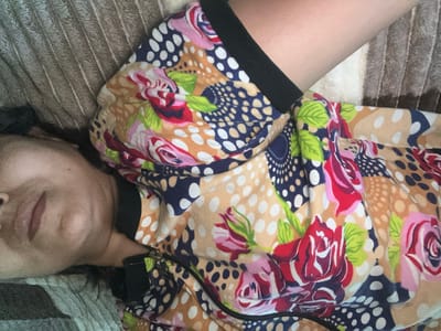 The image shows a person lying down on a textured surface. The person's face is partially visible, with their eyes closed and lips slightly parted, suggesting they are asleep or resting. They are wearing a colorful, patterned garment with a zipper down the front. The pattern features large roses in shades of red, pink, and white, interspersed with polka dots in navy blue, white, and beige. The background surface appears to be a sofa or bed with a ribbed texture, possibly a blanket or upholstery. The lighting in the image is soft, indicating it may be an indoor setting, potentially during the day. There are no visible text elements in the image. The person's arm is also visible, resting against the patterned garment. The overall impression is one of a quiet, relaxed moment.