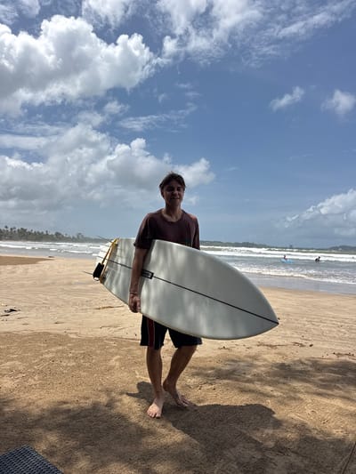 A young man stands on a sandy beach, holding a white surfboard. He is wearing a dark maroon t-shirt and dark shorts with a red stripe. His left hand grips the surfboard, and his right arm is bent, with his hand resting on the board. He is looking towards the camera with a slight smile. The surfboard is angled across his body, with its nose pointing upwards.
The scene is a beach with the ocean and waves in the background. There are other surfers in the water, one on a blue board and another on a dark board. The sky is a vibrant blue with scattered white clouds, suggesting a sunny day. Palm trees are visible in the distance along the coastline, and the sand is a light brown color with shadows cast by the sun. The overall impression is one of a relaxed and enjoyable day at the beach.
The time of day appears to be daytime, likely late morning or afternoon, given the bright sunlight and shadows. The weather is pleasant and sunny. The location cues like palm trees and the type of waves suggest a tropical or subtropical coastal area.
There is some visible text on the surfboard, but it is too small to read clearly. However, it appears to be a brand name or model number. The orientation of the image is portrait, as the man is standing and holding the board. The background is slightly blurred, drawing focus to the man and the surfboard.