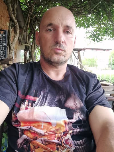 A bald, middle-aged man with light stubble on his chin and upper lip is featured in a close-up, casual self-portrait. He has green or hazel eyes and a neutral, slightly serious expression as he looks directly at the camera.
He is wearing a dark, short-sleeved t-shirt made of a textured fabric. The shirt displays a prominent graphic design: a large, stylized skull at the bottom, with a frothy, overflowing substance resembling beer foam emerging from its top. Above this, a vague cityscape or structural elements are visible, rendered in a palette of orange, white, red, and dark hues.
The man is seated outdoors, under a natural canopy of dense green foliage and thick, gnarled woody vines or branches. To his left, behind him, a reddish-brown brick wall is partially visible, along with a dark metal grate or window structure. The vines are heavily intertwined, covering parts of the wall. To his right, the background is brighter, revealing more distant green trees and a light-colored building with a reddish roof, indicating a residential or semi-rural setting. Indistinct objects, including a round, flat stone-like item, are present on a surface to his right.
The lighting suggests it is daytime, with soft, diffused light indicating shade or an overcast sky. The scene depicts a casual outdoor space, likely a patio or garden area of a building in Obrochishte, Bulgaria. No discernible text is visible in the image.