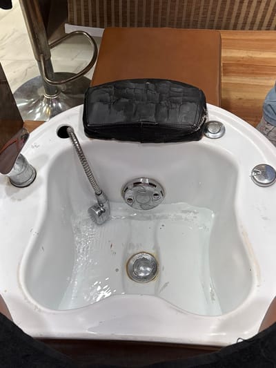 The image depicts a first-person view of a pedicure station within what appears to be a salon or spa in South Dumdum, India. A white, elongated, ceramic or acrylic foot bath is centrally positioned, partially filled with clear water flowing from a flexible chrome showerhead on the left side. A stationary chrome faucet is visible to the far left. Embedded in the rim of the foot bath are multiple circular chrome controls. Resting on the upper back rim of the foot bath is a severely worn and cracked black faux leather cushion, likely serving as a footrest or part of a seating arrangement. The lower sections of a person's legs, covered in dark fabric, are visible at the bottom of the frame, indicating the perspective of a client. In the background, to the left, a portion of a silver metallic adjustable bar stool with a circular base is visible. To the right and behind the foot bath, a brown upholstered square stool or seat is present, set against a wall featuring vertical wooden paneling. The foot bath itself shows some minor discoloration or scuff marks consistent with regular use.