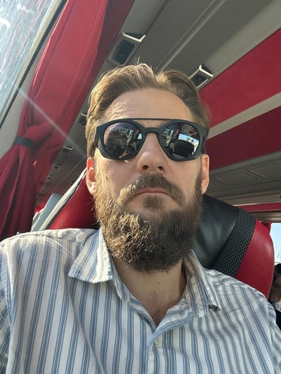 A man with light brown hair and a full beard, wearing sunglasses and a blue and white striped shirt, sits in a vehicle. The interior of the vehicle features red curtains on the side and black and red seating. The man is looking directly at the camera. The scene appears to be set within a bus or similar mode of transportation. The location is Döşemealtı, Turkey.