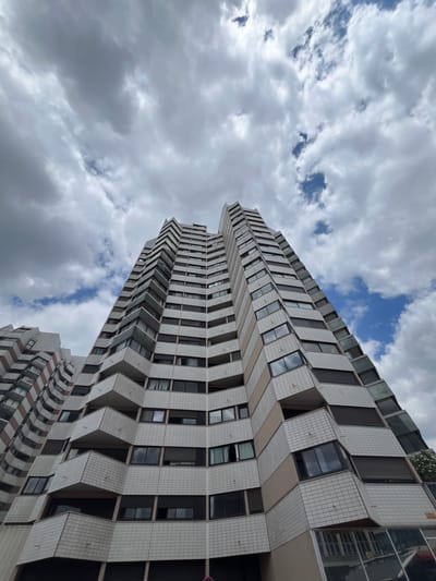 This is a low-angle, full shot of a modern, multi-story apartment building located in Créteil, France. The building features a distinctive zigzagging facade with a repetitive pattern of white tiled sections and beige horizontal bands. Numerous balconies, some recessed and some protruding, are visible, interspersed with windows, many of which are dark, suggesting they are either closed or have dark blinds. The architecture is angular and geometric, giving the building a somewhat futuristic appearance.
The sky above is filled with a mix of dramatic, dark grey clouds and patches of bright blue sky with scattered white cumulus clouds. The lighting suggests it is daytime, but the cloudy conditions create a somewhat overcast atmosphere. No people are visible in the image. The perspective emphasizes the scale and height of the building as it stretches upwards towards the sky. The overall scene captures an urban architectural landscape under a dynamic sky.