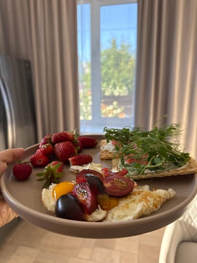 A plate of food is presented against a backdrop of a window with sheer curtains. The plate contains strawberries, a fried egg, sliced tomatoes, a black plum, and two crackers topped with cream cheese and sprouts. The setting appears to be indoors, possibly a home or apartment in Oktyabrsky, Russia, given the provided location information. The light filtering through the window suggests it is daytime.
