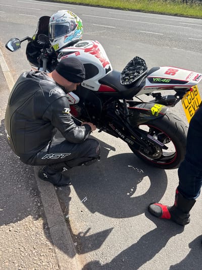 In the city of Much Dewchurch, United Kingdom, a person wearing a black leather motorcycle suit is crouching beside a white and red motorcycle, appearing to be performing maintenance. The motorcycle has a white helmet with a graphic design resting on its fuel tank. The background shows a paved road and a grassy verge, suggesting an outdoor setting on the side of a road.