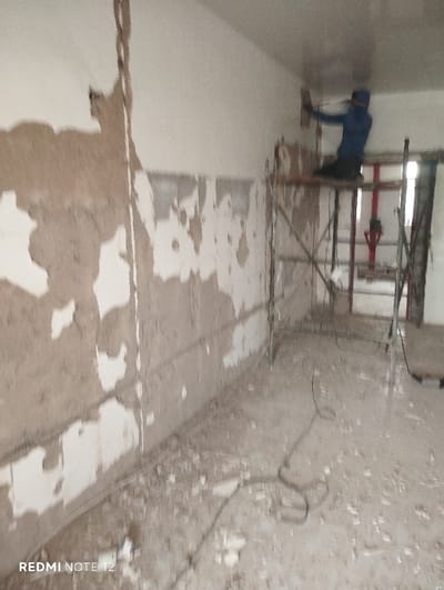 An interior space under renovation. A worker on scaffolding smooths a ceiling. Walls are stripped, revealing layers of material. Debris litters the floor. A red pipe fixture is visible in the background. The space is in a state of disrepair, awaiting further work.