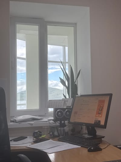 The image depicts a desk in an office environment with a window overlooking a landscape. The desk is cluttered with various items including a computer monitor, keyboard, mouse, papers, cell phones, and a desk chair. A plant is placed on the windowsill, and speakers are positioned near the plant. The computer monitor displays a calendar with dates and weather information. The window shows a view of hills and a cloudy sky. The office appears to be located in the city of Yakty-Kul, Russia.