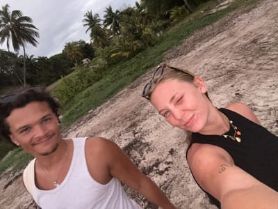Two people are taking a selfie on a beach. A man with curly hair and a white tank top stands to the left. He is smiling. To his right is a woman with blonde hair wearing a black tank top and sunglasses resting on her head. She is winking. They are on a sandy beach with palm trees and vegetation in the background. The sky is overcast. The location is likely in a tropical area. Based on the prompt it should be at Sainte-Anne, France.