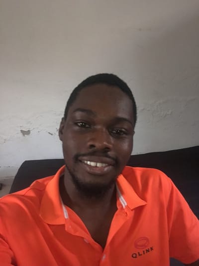 A male individual, appearing to be in his twenties or thirties with dark skin and short dark hair, is depicted in a close-up, chest-up portrait. He has a dark beard and mustache, and is smiling, revealing his upper teeth. The subject is wearing an orange short-sleeved polo shirt with a white inner collar lining visible. A dark grey logo and text, "QLINK", is present on the left chest area of the shirt. He is looking directly into the camera. The background consists of a light-colored, possibly off-white, interior wall that shows signs of wear and discoloration, including peeling paint or plaster on the left side of the frame. A dark, indistinct surface, possibly a chair or sofa back, is visible behind the subject. The scene is located in Katcha, Nigeria.