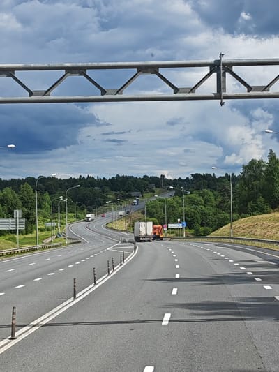 The image depicts a highway scene in Mironegi, Russia. A wide, grey asphalt highway stretches into the distance, curving to the right. The road is marked with white dashed lines indicating lanes. Traffic posts, which are brown and cylindrical with reflective stripes, line the right shoulder of the road. In the foreground, a white truck with an orange rear section is driving on the highway. Further down the road, other vehicles, including trucks and cars, are visible.
The highway is bordered by lush green trees and vegetation on both sides, with a gentle slope rising on the right. The sky above is dramatic, filled with dark, heavy storm clouds contrasted with patches of lighter sky. Overhead, a large metallic structure, likely a sign gantry, spans across the highway. Streetlights are positioned along the sides of the road. The overall impression is of a busy, open road under a turbulent sky in a rural or semi-rural setting.