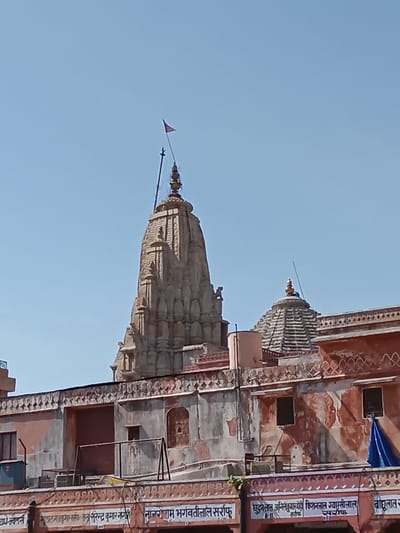 The image shows a building with two temple spires against a clear blue sky. The building's exterior is decorated with intricate patterns and inscriptions in Hindi. The lower part of the building has signs with names and the word "Sarraf," suggesting it could be related to a jewelry business. Based on architectural style and text, this appears to be a structure in Jaipur, India.