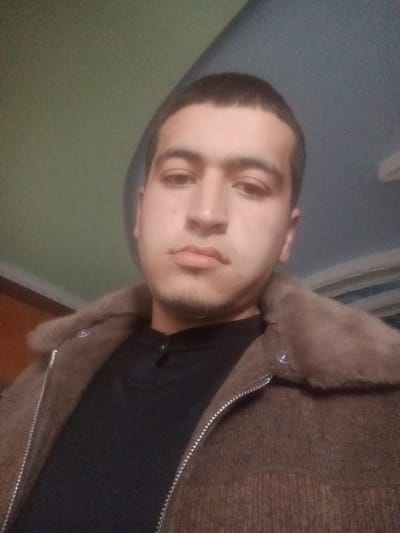 A young adult male is depicted in a head-and-shoulders portrait, likely a selfie, taken indoors. He has short dark hair, dark eyes, and minimal stubble on his chin and upper lip, with a neutral facial expression. He wears a black long-sleeved shirt with a mandarin collar and three visible buttons on the placket. Over the shirt, he wears a brown jacket, possibly of corduroy or ribbed material, featuring a prominent brown faux fur or fur collar and a silver-toned zipper. A single snap button is visible on the jacket's right lapel area. The indoor background consists of a light teal or green colored wall/ceiling segment in the upper portion and a darker, indistinct area on the left. A light-colored fixture is partially visible on the upper right side of the frame. The scene is located indoors, in Kasan, Uzbekistan.