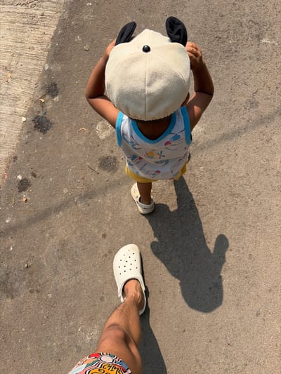 The image shows a child from a top-down perspective, wearing a white cap with black ears and a light-colored t-shirt with a blue trim. The child is standing on a concrete surface, which appears to be a sidewalk or pavement. To the right of the child, an adult's leg is visible, wearing a white Croc-style sandal and colorful patterned shorts. A shadow of a person is cast on the ground to the right of the child, indicating it is daytime with strong sunlight, likely mid-morning or afternoon. The concrete surface shows some discoloration and debris. The overall scene suggests a casual outdoor setting in Denasri Kulon, Indonesia. No text is visible in the image.