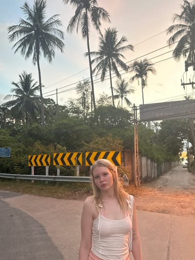 A young Caucasian woman with blonde hair stands facing the camera on a paved road, with her body angled slightly to the right. She is wearing a white tank top over a pink bra and pink shorts. Behind her, palm trees line the left side of the road, and a weathered fence runs along the right. A street sign with Thai writing is visible above the fence, and a few cars are parked further down the road. The sky is a pale orange and yellow, suggesting either sunrise or sunset. There is a yellow and black chevron sign on the left side of the road. The woman's expression is neutral. The overall scene appears to be in a tropical or subtropical location, possibly in or near Tong Yang, Thailand, given the presence of palm trees and Thai signage.
