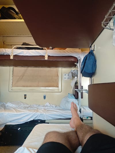 The image depicts the interior of a passenger train sleeping compartment, specifically located within or passing through the city of Druzhinino, Russia. The perspective is from a lower bunk, looking towards the foot of the compartment. In the immediate foreground, the bare feet and lower legs (clad in black shorts) of an individual are visible, resting on a white-linened bunk.
Above this, another lower bunk is prepared with white sheets, a pillow, and a folded blanket. A black hard-shell suitcase with wheels is stowed underneath this bunk. Centered on the wall beyond the lower bunk is a window with a light-colored, opaque shade fully drawn. To the right of the window, a light-colored wall features two small metal hooks. Further right, a vertical metal pole supports a brown shelf, a wire basket containing items (possibly laundry or personal effects), and a blue backpack hanging from a hook. Two small square white stickers with black markings are affixed to the wall above the shelf.
An upper bunk, also made with white linen and a headrest, is visible above the lower bunk. A second individual is lying prone on this upper bunk, with their head and arm partially visible. Above the upper bunk, a brown overhead shelf holds a black backpack. A large, dark brown panel extends from the ceiling, partially obscuring the upper section of the image frame. The compartment walls are a light beige or cream color.