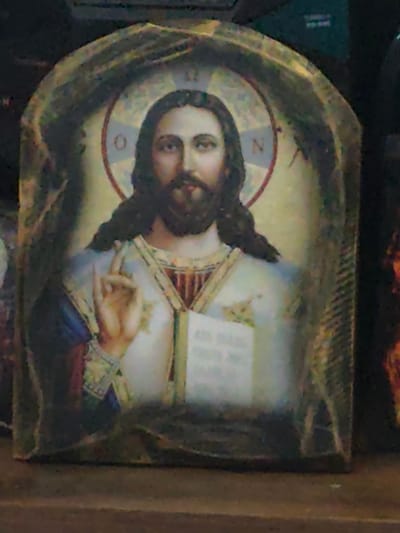 An arched icon depicting Jesus Christ is presented centrally in a dimly lit indoor setting. Christ is shown from the waist up, facing forward with a direct gaze, featuring long dark hair and a dark beard. A golden cruciform halo encircles his head, displaying the Greek letters 'O' on the left and 'N' on the right within its circular boundary. His right hand is raised in a gesture of blessing, with the thumb, ring, and pinky fingers joined, and the index and middle fingers extended. His left hand holds an open book, presumably a Gospel, on which faint, illegible text is visible on the pages. Christ is attired in a white tunic, covered by a blue mantle draped over his left shoulder, and gold-embroidered vestments around his neck and chest. The icon itself appears to be a printed image mounted on a solid material, possibly wood, featuring a textured, dark brown border. The object rests on a flat, dark brown surface, consistent with a shelf or table. The background is dark and out of focus. This item was observed in the city of Lom, Bulgaria.