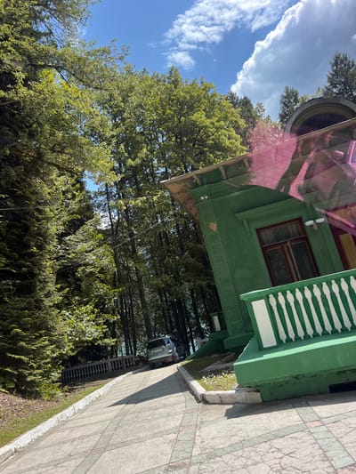 This media file depicts an outdoor scene with a focus on a green, two-story house. The house features a red-trimmed window and a distinctive white-railed porch or balcony. A pink-tinted, transparent canopy extends from the house, reflecting the sky and trees.
Surrounding the house is lush green foliage, including large trees with dense canopies and smaller bushes. The ground in front of the house appears to be a paved walkway with a pattern of grey and pinkish tiles. A small patch of grass with white flowers is visible along the edge of the walkway and house.
In the background, tall trees are visible, and a dark-colored SUV is parked on a road or driveway, partially obscured by the trees. The sky is blue with scattered white clouds, suggesting it is daytime, likely morning or afternoon, with fair weather. The overall atmosphere is peaceful and natural. There are no people visible in the image. The image is taken from a slightly elevated perspective, looking downwards and to the side.
There is no discernible text visible in the image. The scene appears to be set in a location with abundant vegetation, possibly a residential area or a park. The orientation of the image is rotated, so the house and walkway appear to be on a steep incline.