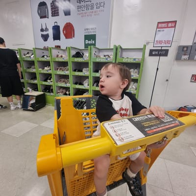 In a retail store in Wonju-si, South Korea, a young child sits in a yellow shopping cart. The child wears a black and white t-shirt and is facing to the right, with their mouth slightly open. The shopping cart has a yellow handle and a black interior grid. Attached to the side of the cart is a warning sign with Korean text and icons depicting children. The background shows rows of shelves filled with footwear, primarily branded with "crocs." Some of the footwear is displayed in green boxes. A large promotional poster hangs on the wall behind the shelves, featuring images of clothing and a suitcase, with Korean text. Another sign with Korean text is visible on the wall above the footwear shelves. A person's legs and feet, wearing white socks and black sneakers, are visible on the left side of the image, standing near the shelves. The floor is tiled with a light gray pattern.