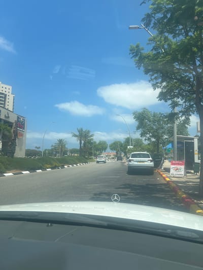The image shows a view from inside a vehicle driving on a road in Or Akiva, Israel. The sky is blue with scattered white clouds. On the left side of the road, there is a modern building with multiple windows and a large sign that says "Amy." Palm trees and green landscaping line the median of the road. Several cars are visible on the road, including a white Skoda with its door open. A white Mercedes-Benz emblem is visible on the hood of the vehicle from which the photo is taken. On the right side of the road, there is a bus stop or shelter with a sign displaying "052-3739002." The street is paved and there is a clear demarcation of the road with a black and white striped curb. Trees with green foliage are visible along the right side of the road.