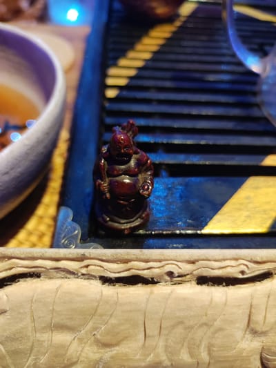 The image displays a static indoor scene focused on a collection of objects. The specific location within Mas, Indonesia, is not discernible from the image content.
**Subjects/Objects:**
1. **Figurine:** A small, dark reddish-brown figurine, approximately 5-7 cm tall, depicting a plump, smiling figure consistent with a Laughing Buddha (Budai or Hotei). It stands upright and is the central focal point.
2. **Tray:** The figurine rests on a dark, possibly black or dark blue, rectangular tray featuring a grid or slatted pattern. Several horizontal elements within this pattern are accented with a reflective gold or yellow finish. A small, translucent, molded plastic or glass decorative piece with an ornate scroll pattern is visible on the tray, immediately to the lower left of the figurine.
3. **Bowl:** To the left, partially visible and significantly out of focus, is the upper rim and interior of a light-colored (possibly white or pale lavender) ceramic bowl. It contains a warm-toned liquid, suggesting a beverage like tea.
4. **Carved Wood:** The bottom section of the image features a light-colored, possibly unfinished, wooden object with intricate, layered horizontal carvings that create a wavy, undulating texture. This object serves as a foreground element.
5. **Glassware:** To the far right, a heavily blurred, transparent object, likely a glass or plastic handle from a cup or teapot, is vaguely visible.
**Environment/Lighting:**
The scene is illuminated by artificial light, contributing to a warm, intimate atmosphere. A distinct, bright blue light source or reflection is prominent in the upper left background, casting a bluish hue onto the dark tray and creating bokeh effects in the background. No individuals are present in the frame, and no dynamic actions or interactions are depicted.