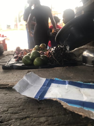 The image shows a street vendor selling fruit in Umudike, Nigeria, located at latitude 5.481 and longitude 7.546. The vendor's stand is located on a wooden plank and consists of a variety of fruits including green mangoes and palm fruits. The vendor is obscured from view, but several customers are visible, some with their hands outstretched, presumably towards the fruit. The foreground consists of a close-up of the fruit stand, with the remainder of the scene out of focus. To the right of the fruit stand is a piece of cardboard with blue and white stripes. There is an empty space to the left of the fruit stand that reveals the street beyond. The scene appears to be lit by sunlight from above.