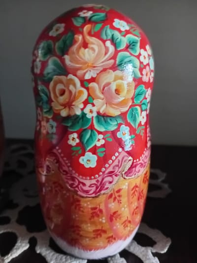 A close-up, eye-level shot shows a brightly painted Matryoshka doll. The doll is primarily red with various floral designs, including large peach-colored roses and smaller white and blue flowers with green leaves. The bottom portion of the doll transitions to an orange and yellow gradient, adorned with smaller red leaf motifs. The doll rests on a dark surface, partially covered by a white, lace-like mat. The background is a blurred gradient of gray and dark brown. The image is well-lit with a soft light source creating subtle shadows. The doll is positioned slightly off-center to the left. There is no indication of the city of Kherson, Ukraine, in the image.