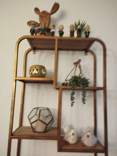 The image shows a close-up view of a tall, gold-colored, geometric shelving unit against a white wall. The shelving unit is decorated with various items. On the top shelf, there are several small figurines of people wearing suits and ties, some with glasses, and a larger wooden reindeer-like decoration at the very top. To the right of the figurines is a small potted plant with spiky green leaves. The second shelf from the top on the left side holds a golden, diamond-patterned candle holder. The corresponding shelf on the right holds a triangular glass terrarium with a hanging succulent plant. The third shelf from the top on the left contains a hexagonal glass and metal geometric lantern with a thick white candle inside. The bottom shelf features two small white owl figurines. The overall impression is of a neatly arranged decorative display. The image does not contain any information about the city of Pardes Hana-Karkur, Israel.