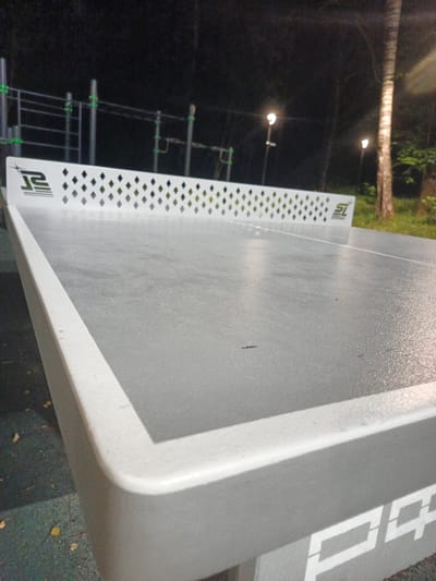 A close-up, low-angle shot of a grey outdoor ping pong table at night. The table has a white border and a white net with diamond-shaped cutouts. The logos of "JS" and "SL" are visible on the net. In the background, there are streetlights, trees, and outdoor fitness equipment. The ground appears to be dark asphalt or rubber surfacing with fallen leaves. The overall setting suggests a public park or recreational area.