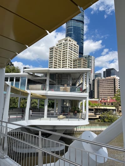 The image, viewed in landscape orientation, presents an urban waterfront scene in Brisbane City, Australia, under a clear blue sky with scattered white clouds. In the immediate foreground, a complex, multi-tiered white architectural structure with glass panels and metal railings occupies the lower half, possibly serving as public viewing platforms or open-air dining areas. Several individuals are visible within these structures; one person is seated at a table on a mid-level platform, and others are seen standing or sitting elsewhere.

To the right, a large, sculptural white structure with prominent railings extends into a body of water, likely a river. This structure appears to be part of a pier, ferry terminal, or a significant waterfront feature.

The background showcases a dense cityscape. On the left, a striking, tall skyscraper with curved blue glass facades dominates the skyline. Other high-rise buildings, varying in architectural style and color from beige to grey, fill the mid-ground. Faint text "PARK BLDG" is discernible on one background building, and "Queens Wha" (likely Queens Wharf) is partially visible on another brown building, consistent with the Brisbane location. The overall impression is one of a bright, sunny day in a modern, active city environment.