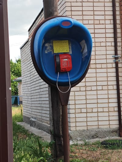 A weathered, blue and brown public payphone booth stands in front of a light-colored brick building. The booth's blue interior houses a red, coin-operated telephone with a keypad and a small screen. A yellow note with handwritten text in a foreign language is affixed to the inside of the booth. The phone's coiled cord is visible, and a metal pipe, likely part of the booth's support structure, extends downwards. The building is made of light-colored bricks laid in a stretcher bond pattern, with a grey foundation. Green grass and some weeds grow around the base of the booth and the building. The scene appears to be outdoors during the daytime, with a cloudy sky visible. The overall impression is one of disuse and neglect. The location is Krutinskiy, Russia.