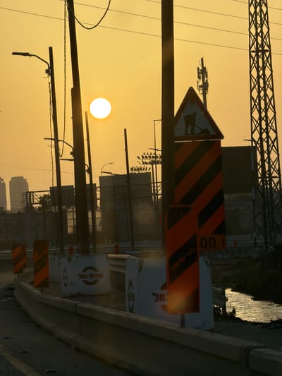 The image captures an urban or industrial landscape at either sunrise or sunset, characterized by warm golden and orange hues dominating the sky. The scene, located in Azor, Israel, features prominent silhouettes of structures against the bright sky.
In the upper portion of the frame, a large, intricate metal lattice structure, possibly part of a bridge or power transmission tower, stretches across. Below it, several thick, dark horizontal poles with attached wires and lighting fixtures traverse the sky. A bright, circular white sun is visible in the middle-left, casting long shadows and creating a striking contrast with the dark foreground elements.
The foreground and midground contain several construction or road-related objects. A triangular warning sign with a black silhouette of a person shoveling dirt is visible, indicating road work. Nearby, multiple rectangular signs with distinctive black and orange diagonal stripes are present, commonly used for construction zone warnings. One such sign partially reveals the numbers "00 1". On the right side, a series of white barriers or drums are aligned. Hebrew text is visible on some of these barriers; one clearly displays "פיבי בראל" (Pibi Barel), suggesting a construction company, and another partially shows "אל" (El).
In the background, dark silhouettes of buildings and other urban structures are visible, hinting at a developed area. To the far right, a body of water, possibly a river or canal, shimmers with the reflection of the low sun. A dark, elevated path or road runs alongside this water body. The overall impression is one of ongoing development or infrastructure within an urban setting, beautifully illuminated by the dawn or dusk light.