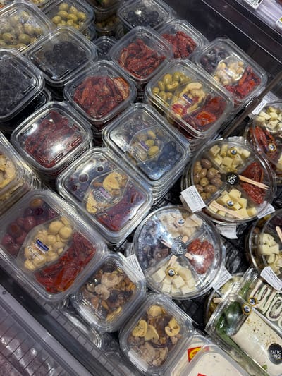 The image shows a refrigerated display case containing various pre-packaged antipasto items, likely located in a supermarket. The items are in clear plastic containers of square and round shapes. The contents include marinated olives (green and black), sun-dried tomatoes, marinated mushrooms, artichoke hearts, cubed cheese, and other appetizers. Some of the round containers are divided into sections containing different components, such as cheese, olives, and sun-dried tomatoes. Labels on the packages indicate brands such as "FATTO da NOI", "PANEBIANCO i freschissimi", "Trisolivo", "Sfizioso", and "Gustoso". Some packages also have labels that read "Superstore Deco" and "POKER SFIZIOSO". Based on the OCR and visual appearance, the location is likely in or near Chianchitta, Italy, as "Superstore Deco" is an Italian supermarket chain.