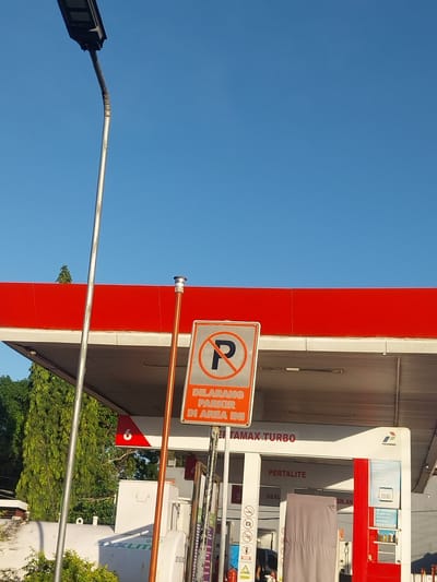 This image captures a street scene at a gas station under a clear blue sky, suggesting a sunny day. The dominant features are the bright red and white structure of the gas station, with its corrugated metal roof. A prominent sign displays a red circle with a diagonal line over a "P" symbol, indicating "DILARANG PARKIR DI AREA INI," which translates to "NO PARKING IN THIS AREA." Below this, the words "PERTAMAX TURBO" and "PERTALITE" are visible, identifying fuel types offered.

To the left, a street lamp with a modern design is partially visible against the sky. In the background, lush green trees provide a natural contrast to the man-made structures. The gas station appears to be operational, with some equipment and signage indicating services. The overall impression is one of a typical day at a well-maintained gas station, with clear visibility and good weather conditions. The presence of Indonesian text suggests the location is likely in Indonesia, potentially on the island of Java given the mention of "Macege." No people are clearly visible in the image.