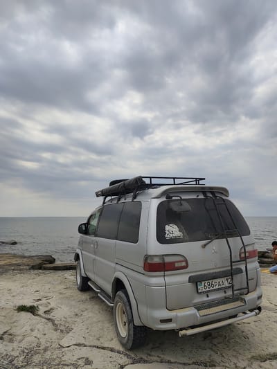 A silver Mitsubishi Delica van is parked on a rocky, flat coastline overlooking the sea. The van has a roof rack with an awning and is facing away from the camera, with its rear visible. A ladder is mounted on the back of the van. A license plate reading "KZ 686PAA 12" is attached to the rear bumper. A sticker on the rear window says "DELICA AKTAU CLUB". The sky is overcast with gray clouds. In the background, to the right, a person is seated on the rocks. The location is identified as Aqshuqyr, Kazakhstan.