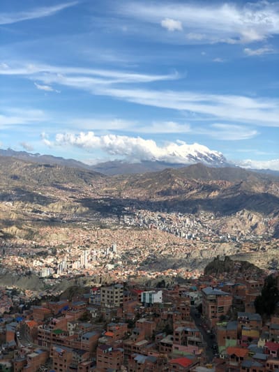 A high-angle view shows a sprawling city nestled along a rugged mountain range. Brick buildings of varying heights tightly pack the urban landscape. A snow-capped mountain peak pierces the clouds in the distance. The sky above is a mix of blue and white. The terrain transitions from barren mountainous slopes to a densely populated cityscape. The city's layout seems organic and sprawling, clinging to the contours of the terrain.