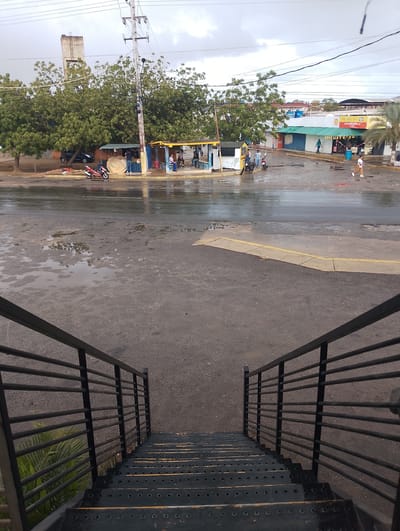 The image captures a street scene in El Espinal, Venezuela, observed from the top of a dark metal staircase with horizontal bar railings. The foreground features the descending staircase, leading to a wet asphalt road characterized by numerous puddles and reflective surfaces, indicating recent rainfall. A yellow-painted curb defines the edge of the road. In the midground, a red motorcycle is parked adjacent to a small, makeshift stall where a man in a dark shirt is standing. To the right, several individuals are seated under a yellow-roofed structure. A black car is partially visible behind dense green trees on the left. Overhead power lines and a prominent utility pole traverse the upper-midground. The background consists of a row of single-story commercial buildings, including an "AUTO PARTS" store with "SHELL" branding, some with closed security shutters. Pedestrians are visible walking on the far side of the street. A tall, slender concrete water tower rises above the trees on the left, under an overcast sky.