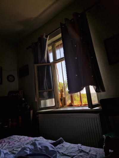 This is an indoor, eye-level, medium shot of a bedroom window in Raduvene, Bulgaria, as illuminated by natural light filtering through the window. The view is from a bed, with a white and pink patterned duvet and a white crumpled sheet in the foreground. A radiator is visible beneath the window. The window itself is old, with a weathered white wooden frame, and is partially open, revealing an exterior scene of green trees and vegetation bathed in warm, late-day sunlight. Two sets of curtains are drawn across the window: one is a sheer white curtain with small stars, and the other is a darker, opaque blue curtain with a scalloped hem. The room is dimly lit, with shadows cast on the walls and ceiling. On the wall to the left, a framed circular object resembling a clock and two other framed pictures are visible. To the left of the window, a dark wooden cabinet or dresser is partially visible, with some items on top.