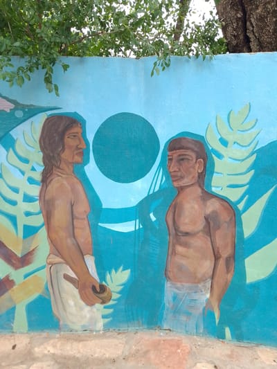 A mural in Villa Hayes, Paraguay, depicts two shirtless indigenous men wearing loincloths. One man on the left is holding a round object, while the other stands with his arms at his sides. The mural's background is light blue with stylized green plant life and a dark blue circle. The mural is partially obscured by tree branches and a tree trunk at the top. It is painted on a wall above a concrete base.
