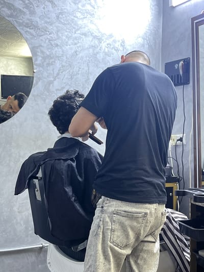 In a dimly lit barber shop in Al Machouar – Stinia, Morocco, a barber with a shaved head and a black t-shirt is meticulously trimming the hair of a client sitting in a black salon chair. The client, wearing a black cape, has curly dark hair and is facing away from the camera. The barber is holding a comb and a razor, actively engaged in the haircut. The shop's walls are textured with a grey, mottled finish. To the left, a large, oval mirror reflects the back of the client's head and a portion of the barber shop. On the right side of the frame, another mirror with a wooden frame is visible. The scene is cluttered with barber shop equipment, including electrical outlets and tools organized on a rack, indicating a functional workspace. The overall atmosphere suggests a typical day at a local barbershop.