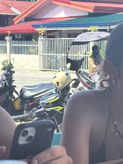This image depicts an outdoor street scene located in Burgos, Philippines, under bright daylight. In the immediate foreground, the right shoulder, ear, and dark hair of a person are visible, adorned with multiple necklaces. This individual holds a black smartphone, featuring a triple-camera setup, in their right hand. A segment of a light blue surface is positioned beneath the phone.

The midground features a paved street with multiple motorcycles and scooters parked alongside. A black and yellow scooter is visible, with a cream-colored helmet, labeled "FOR ACE," resting on its seat. To the right, another person is seated on a black motorcycle equipped with a dark, semi-circular shade umbrella. The rider is dressed in a light brown long-sleeved shirt and gray shorts, with their head completely obscured by a dark balaclava or head covering.

The background comprises residential buildings with distinctive red and blue corrugated metal roofs. White vertical bar fences separate the properties from the street, and small potted plants are visible on some ledges or atop the fences.