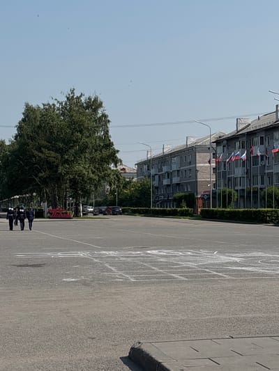 In Belovo, Russia, a sunny day is depicted on a wide, paved area that appears to be a public square or plaza. Three individuals in dark uniforms, possibly ceremonial or law enforcement, walk from left to right across the foreground, their figures silhouetted against the bright pavement. To their right, a large, leafy green tree stands prominently, its branches reaching upwards. Beyond the tree and pavement, a row of multi-story residential buildings with grey facades lines the street. Several flags, likely national or regional, are displayed on poles in front of these buildings. The sky above is a clear, pale blue, with a few distant birds visible. The ground is marked with white chalk lines, suggesting recreational activities or a planned event such as hopscotch or a game. Cars are parked along the street in the middle ground. The overall impression is one of an ordinary day in a Russian city, with civic structures and natural elements coexisting.
