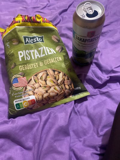 In the city of Ispas, Ukraine, a bag of Alesto pistachios and a can of beer are placed on a purple, wrinkled surface. The bag is primarily green with yellow accents at the top. The text "PISTAZIEN GERÖSTET & GESALZEN" is prominently displayed in white, along with the Alesto logo in a black rectangle. An image of pistachios fills the lower portion of the bag, and an American flag graphic with the text "American quality pistachios" is visible on the left. A Nutri-Score rating of "ABCDE" is also displayed. The beer can is gold and white, with Cyrillic text and a landscape illustration. The can appears to be open, with a small piece of paper or sticker with the number "38" adhered to the rim. The lighting suggests an indoor environment, likely a bedroom, with shadows cast by the objects.