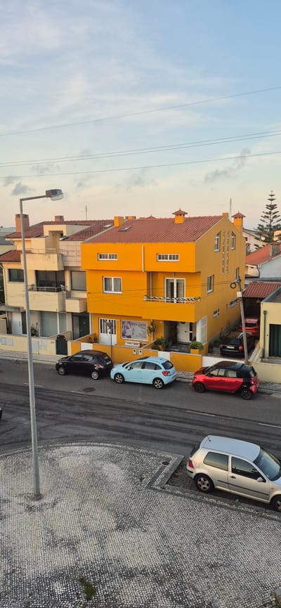 The image captures a street scene in Póvoa de Varzim, Portugal, on a clear day with a light blue sky and scattered clouds. The foreground features a cobblestone sidewalk with a lamppost and the edge of a road marked with tire tracks. Several cars are parked on the street: a silver hatchback, a black hatchback, a light blue hatchback, a red and black smart car, and a red sedan visible further back.
Dominating the midground is a vibrant, two-story, bright orange building with a red-tiled roof. The building has several windows and balconies. A sign with "YOYO LOOK" and an image of a woman with styled hair is visible near the entrance. Adjacent to the orange building is a lighter-colored, multi-story building with a balcony.
In the background, other residential buildings with red-tiled roofs are visible, along with a tall, slender evergreen tree. Overhead, power lines crisscross the sky. The overall impression is of a residential or mixed-use urban environment. There are no people visible in the image.