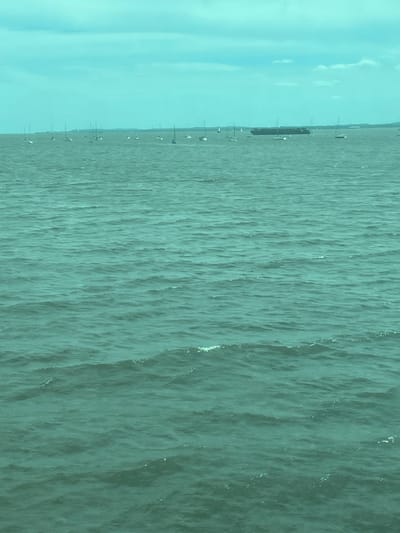 The image depicts an overcast daytime scene of the Raritan Bay or Arthur Kill waterway, viewed from the city of Perth Amboy, New Jersey, United States. The lower two-thirds of the frame are dominated by teal-tinted water exhibiting moderate choppiness and small waves. In the mid-ground, approximately two dozen stationary sailboats are scattered horizontally across the water, identifiable by their visible masts and light-colored hulls. A large, dark-hulled cargo or container ship is also present in the mid-ground, positioned right of center, appearing stationary. The upper third of the image shows a light teal, cloudy sky with diffuse illumination. A faint, indistinct landmass is visible on the distant horizon, particularly towards the right. No people or specific actions are discernible.