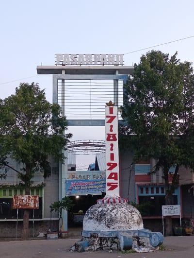 A large sign with the text "PASAR PANJI" in white letters against a blue background is mounted on a tall, blue and white structure. The structure has a white, dome-shaped base that is adorned with red and blue trim. The sign stands in front of the entrance to a building with blue and green walls. There is a large, white sign with text in Indonesian hanging on the wall. The sign is surrounded by green trees, some of which have brown trunks. There are a few people standing in front of the structure and a few parked vehicles in the background. The scene is located in Kecamatan Panji, Indonesia, at coordinates -7.699 latitude and 114.03 longitude. The ground is paved and there is a blue-green color hue over the image.