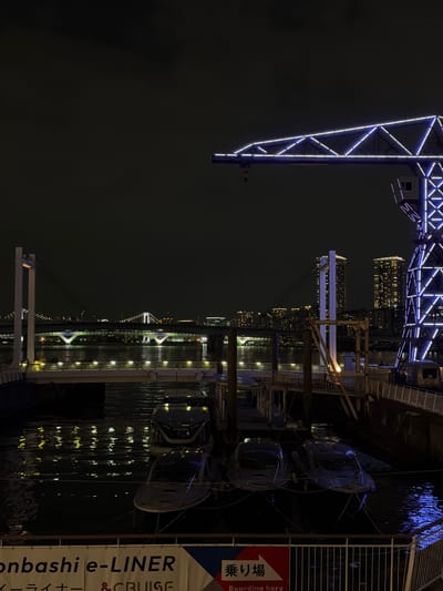 The image depicts a nighttime scene in Koto, Japan, likely near the waterfront. A large, illuminated crane with white and blue lights is prominently featured on the right side of the frame. In the background, a brightly lit bridge spans across a body of water, with city buildings visible beyond it. Several boats are docked in the foreground, their reflections shimmering on the dark water. A sign with Japanese text and the words "e-LINER & CRUISE" is visible at the bottom of the image, suggesting a ferry or cruise terminal. The overall atmosphere is urban and industrial, with the lighting creating a dramatic and slightly futuristic feel. No people are clearly visible in the image.