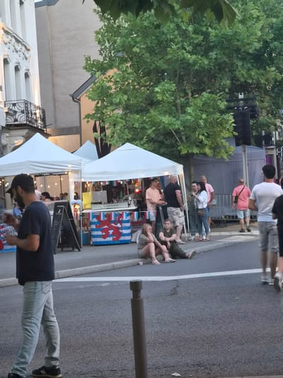 The image depicts an outdoor event in Differdange, Luxembourg. Several people are gathered along a street, some standing and others sitting. Tents are set up, suggesting a market or festival. In the background, trees and buildings are visible, along with what appears to be a stage setup. A flag with a red lion on a blue and white striped background, likely the flag of Luxembourg, is prominently displayed on one of the stalls. Some individuals are interacting with the stalls, possibly purchasing items or enjoying food and drinks. The lighting suggests it is either late afternoon or early evening. The overall atmosphere appears relaxed and casual. No explicit emotions are discernible from the people, but the scene suggests a social gathering. There is also text visible on a sign on one of the tents that appears to say "BOFFERDING".