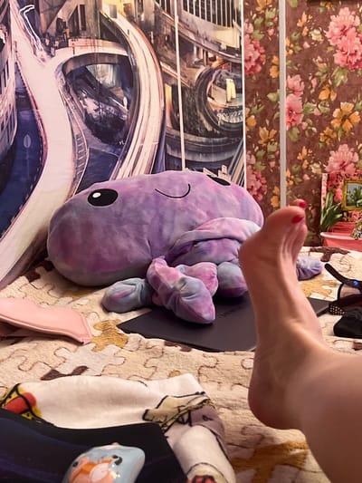 In a dimly lit room, a large, plush purple octopus toy lies on a puzzle-patterned blanket. To the right of the toy, a person's bare foot, with painted red toenails, is propped up, its heel visible. The foot appears to be resting on a dark surface, possibly a laptop or tablet. In the background, a large tapestry or mural depicts an urban cityscape with elevated roadways and buildings. To the right of the cityscape, a floral-patterned wallpaper is visible. A pink pouch is on the blanket to the left of the octopus, and other objects, including a pair of glasses, are scattered around. The overall impression is one of a relaxed, domestic setting. The image does not contain any specific features that would allow for identification of the location as Krasnoe-na-Volge, Russia.